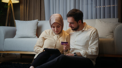 Young married couple leaning against the sofa sitting on floor at home at night, using a tablet...