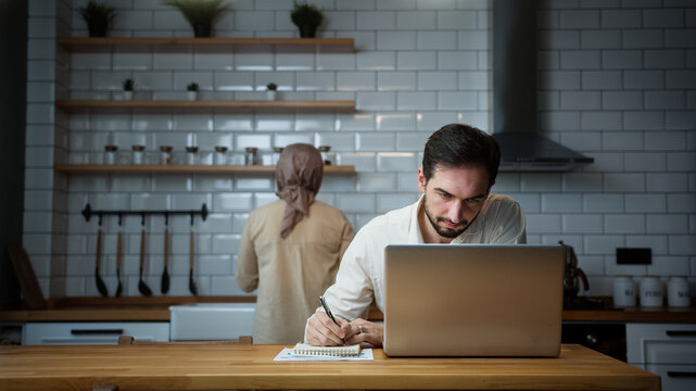 Handsome man is working on his laptop in the kitchen at home while his wife working in kitchen	