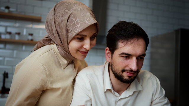 Young man is working and typing on his laptop in the kitchen when his wife comes round from behind and hug	
