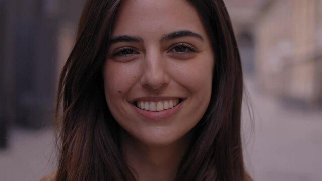 Close up portrait joyful beautiful young Caucasian gen z woman happy smiling on city street. Female people with nice expression looking at camera in open air. Spanish girl posing for picture outdoor