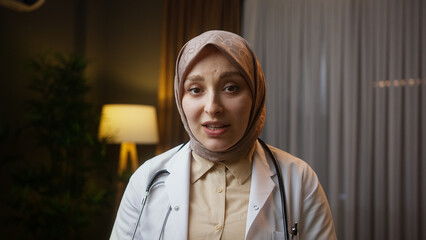 Young female doctor in headscarf wears white coat looking at camera making conference video call....