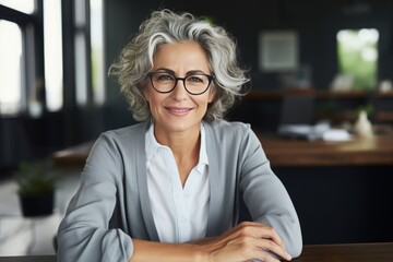 Beautiful middle aged woman, gray hair wearing glasses, sitting at her desk smiling, business woman, healthy
