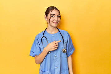 Young nurse in a yellow studio smiling and pointing aside, showing something at blank space.