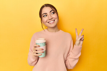 Young caucasian woman holding a takeaway coffee cup joyful and carefree showing a peace symbol with fingers.