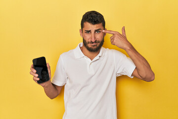 Young Hispanic man shows phone screen showing a disappointment gesture with forefinger.