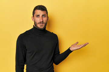Young Hispanic man on yellow background impressed holding copy space on palm.