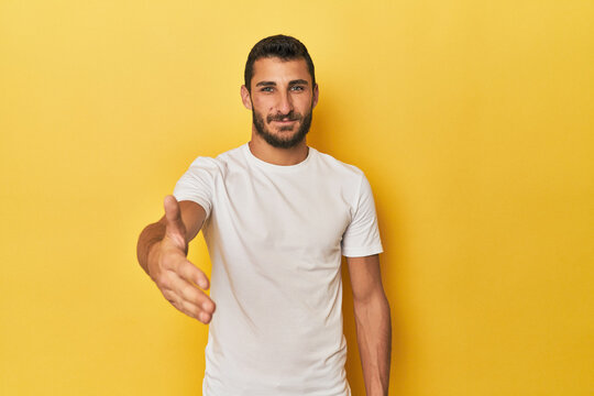 Young Hispanic man on yellow background stretching hand at camera in greeting gesture.