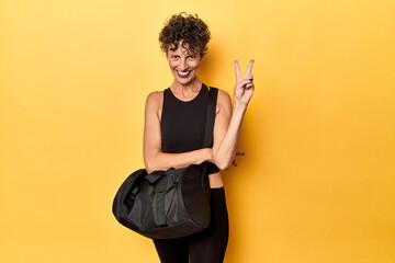 Sportswoman with gym backpack on yellow studio showing number two with fingers.