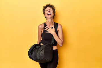 Sportswoman with gym backpack on yellow studio laughs out loudly keeping hand on chest.