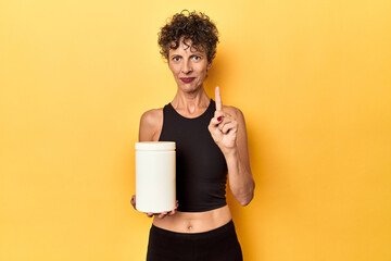 MIddle aged athlete woman holding protein supplement on yellow showing number one with finger.