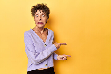 Mid-aged caucasian woman on vibrant yellow shocked pointing with index fingers to a copy space.