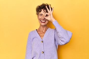 Mid-aged caucasian woman on vibrant yellow excited keeping ok gesture on eye.