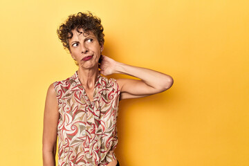 Mid-aged caucasian woman on vibrant yellow touching back of head, thinking and making a choice.
