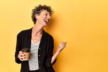 Businesswoman holding takeaway coffee on yellow points with thumb finger away, laughing and carefree.