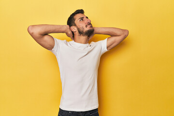 Young Hispanic man on yellow background feeling confident, with hands behind the head.