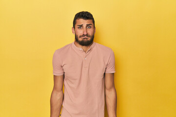 Young Hispanic man on yellow background shrugs shoulders and open eyes confused.