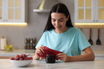 Woman packing jar of jam into beeswax food wrap at table in kitchen