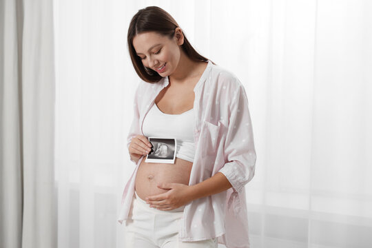 Beautiful pregnant woman with ultrasound picture of baby near window indoors