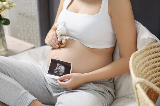 Pregnant woman with ultrasound picture of baby and bunny toy sitting on bed indoors, closeup