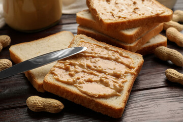 Delicious toasts with peanut butter, nuts and knife on dark wooden table, closeup