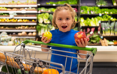 Kid with apple and shopping cart at grocery store. Healthy food for kids. Portrait of smiling...