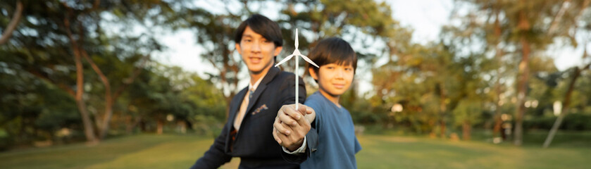 Asian boy and businessman hold wind turbine model together as corporate social responsible to make greener by eco alternative energy for sustainable future generation. Panorama blurred background.Gyre
