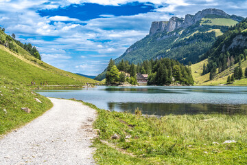 Fototapeta na wymiar Panoramic view of tourists walking along an alpine lake in a green valley with a mountain huts in the background on a sunny day. Seealpsee, Säntis, Wasserauen, Appenzell, Switzerland.