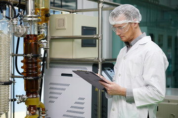 scientist writing on clipboard and looking cannabis extraction machines in testing lab