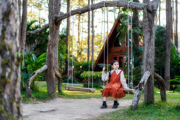 Asian child travel or kid girl sitting relax on wooden swing in nature trees garden or forest and A-frame house background for holiday vacation and tourist happy in resort at Doi Bo Luang Forest Park