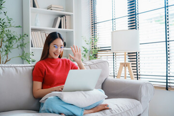 Fototapeta premium Happy young Asian woman wearing eyeglasses using laptop while seated on couch at home