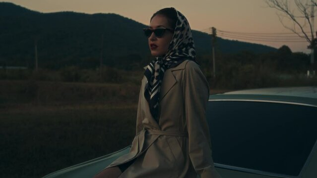 Attractive young elegant woman in a retro outfit sits on the hood of her car. 