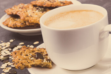 Cup of coffee with milk and fresh baked oatmeal cookies with honey and healthy seeds on white plate. Delicious crunchy dessert