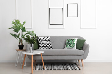 Cozy sofa with cushions and houseplants on tables near white wall