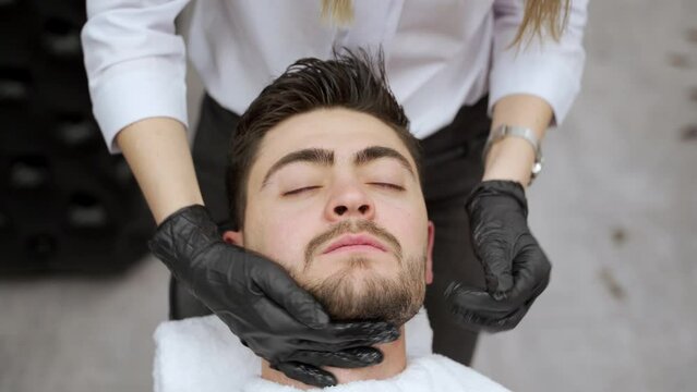 Barber gives relaxing neck face massage to male client lying in chair with eyes closed. Professional wears black gloves for skincare routine in barbershop setting, enhancing client comfort.