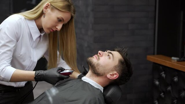 Professional barber gives stylish haircut to male client. Woman expert trims beard, uses clippers in modern shop. Grooming session, mens fashion, hairstyling techniques, salon interior.