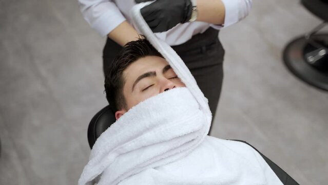 Barber wraps hot towel on customers face for shave prep in salon. Male client relaxing, barber shop facial treatment before beard grooming. Traditional wet shave concept, spa for men.
