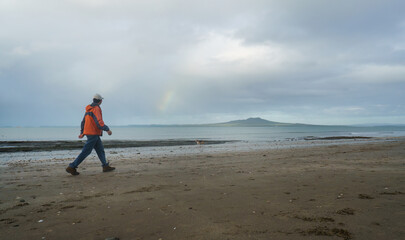 Man walking on the beach. Rainbows over Rangitoto Island in the background. Auckland.