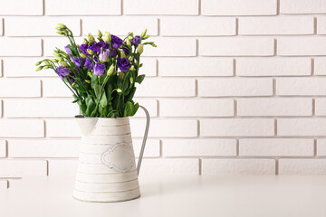 Jug with eustoma flowers on table near white brick wall