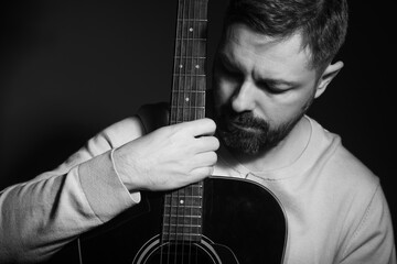 Black and white portrait of a caucasian man holding a guitar in his lap. He is in his 40s and is wearing a white sweater. He has brown hair and a beard. The photo was taken in a studio. - obrazy, fototapety, plakaty