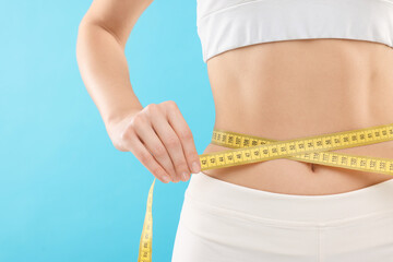 Slim woman measuring waist with tape on light blue background, closeup. Weight loss