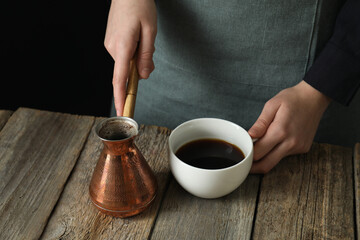 Turkish coffee. Woman with cezve and cup of freshly brewed beverage at wooden table against black background, closeup