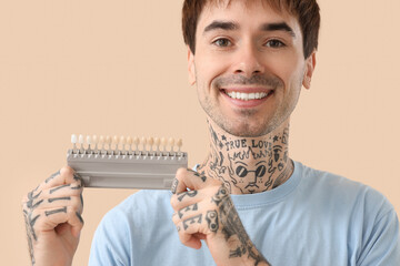 Young tattooed man with teeth color samples on beige background, closeup