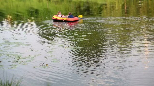 Boy and girl float in inflatable boat with oars along pond.