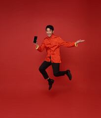 Fototapeta na wymiar Happy Asian man wearing traditional dress jumping and showing mobile phone blank screen isolated on red background.