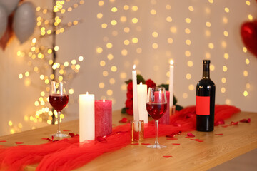 Bottle and glasses of wine, burning candles with decor on table against blurred lights. Valentine's Day celebration at home