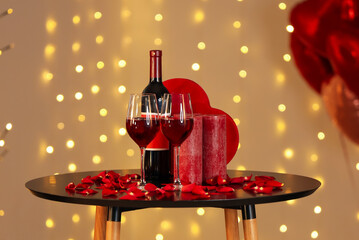 Bottle of wine with gift, candles and rose petals on coffee table against blurred lights....