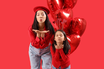 Young Asian woman and her little daughter with heart-shaped air balloons blowing kisses on red background