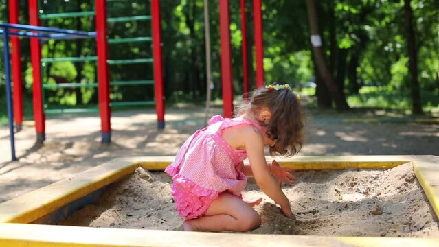 Little girl in a pink dress is digging hole on sandbox.