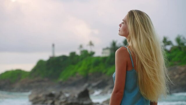 Blonde woman in blue dress strolls along tropical beach, admires lighthouse amid lush nature. Serene seaside walk, soothing landscape, mental wellness backdrop.
