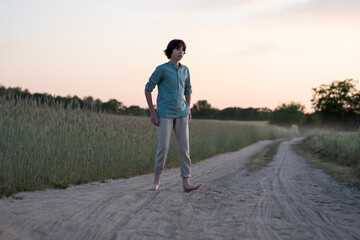 A barefoot teenager in comfortable clothes stands on a dirt road.